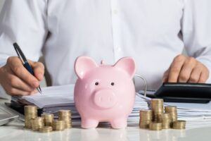 Mann sitzt mit Sparschwein, Münzstapel und Taschenrechner am Tisch und rechnet Geldanlagen durch.