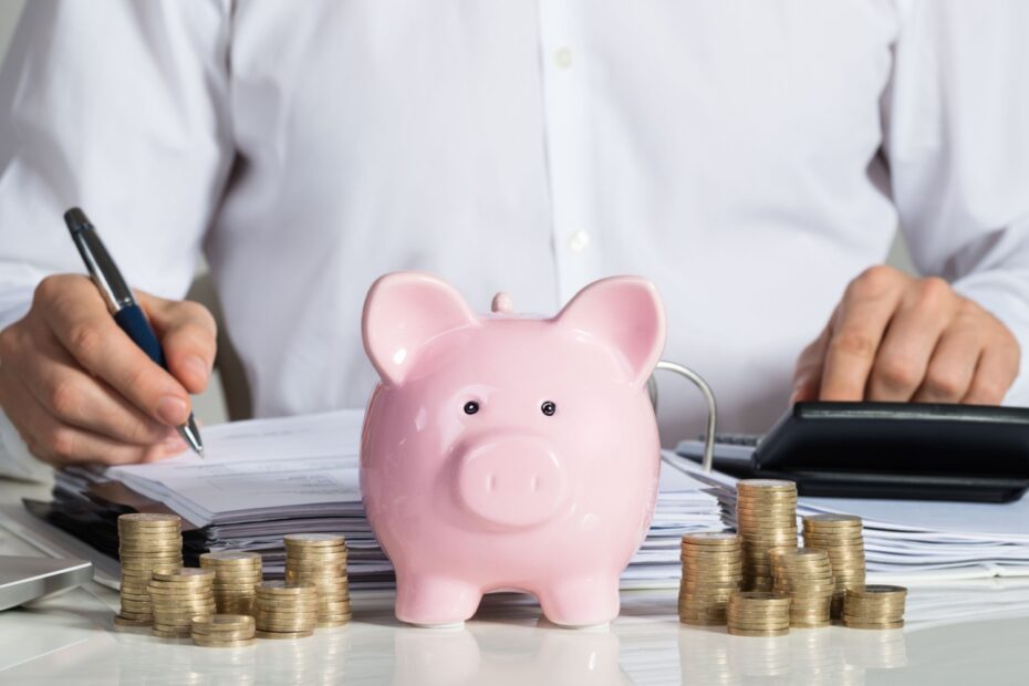 Mann sitzt mit Sparschwein, Münzstapel und Taschenrechner am Tisch und rechnet Geldanlagen durch.